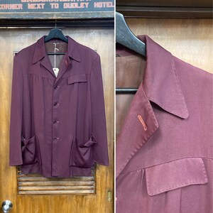 Puede incluir: Chaqueta vintage de color burdeos con cuello con solapas, cuatro botones y dos bolsillos delanteros grandes. La chaqueta está colgada de una percha negra. Un primer plano revela los detalles de la tela y las costuras. La chaqueta está colgada frente a una puerta de madera.
