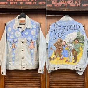 Könnte beinhalten: Eine hellblaue Jeansjacke mit einem bemalten Design auf der Rückseite. Das Design zeigt die Figuren aus dem Zauberer von Oz, darunter Dorothy, der Blechmann, der Feige Löwe und der Vogelscheuche. Der Text "The Wizard of Oz" ist in großen, fetten Buchstaben über den Figuren geschrieben.