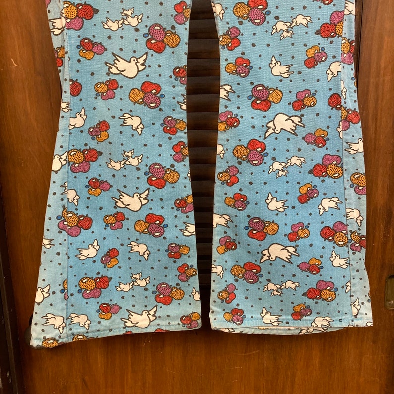 May include: A pair of blue flared jeans with a white, red, and yellow floral pattern. The pattern features doves and clusters of fruit.