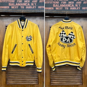 May include: Yellow varsity jacket with black and white striped ribbing. The jacket has a black snap button closure and two black zippered pockets. The back of the jacket has a black and white embroidered logo that says "Kemac D-A Racing Division".