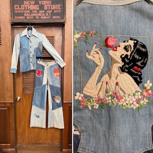 May include: A denim jacket and pants set with patchwork details. The jacket has a light blue denim base with darker blue denim panels. The pants are a mix of light and dark blue denim with various embroidered patches, including a red apple, a flower, and a woman's face.