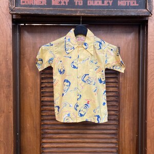 May include: A yellow short-sleeved shirt with a colorful print of faces and musical instruments. The shirt has a button-up front and a collar.