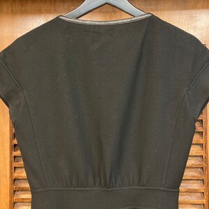 Puede incluir: Un vestido negro de manga corta con un ribete negro alrededor del escote. El vestido tiene un corpi&ntilde;o ajustado y una falda ligeramente acampanada.