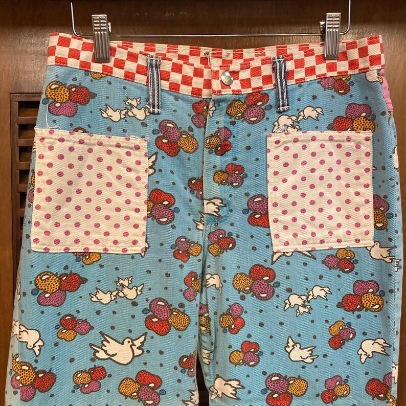 May include: Blue denim jeans with a patchwork design. The jeans have a red and white checkered waistband and two pockets with pink and white polka dot fabric. The main fabric features a pattern of colorful balloons and white doves.
