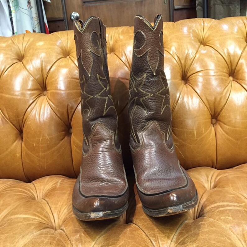 K&ouml;nnte beinhalten: Ein Paar braune Leder-Cowboystiefel mit aufwendigen N&auml;hten und spitz zulaufenden Zehen. Die Stiefel stehen auf einem braunen Ledersofa.