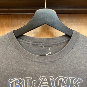 May include: A dark gray t-shirt with the word "BLACK" printed in white and blue on the front. The shirt is hanging on a black hanger.