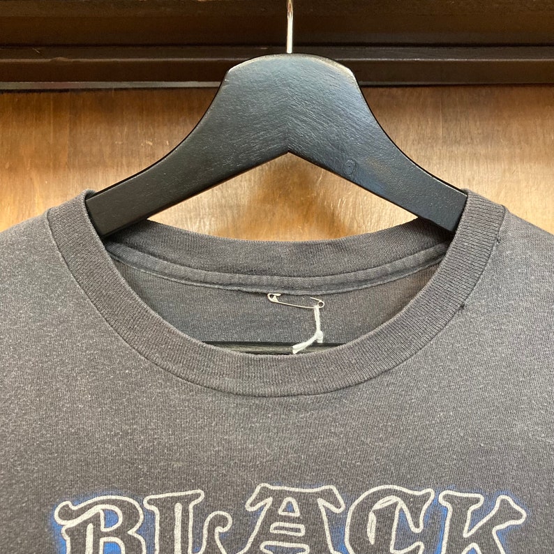 May include: A dark gray t-shirt with the word "BLACK" printed in white and blue on the front. The shirt is hanging on a black hanger.