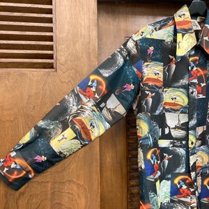 May include: A long-sleeved button-up shirt with a black background and a colorful pattern of cartoon characters and scenes. The characters are in a variety of poses, including flying, swimming, and riding in boats. The scenes are set in a variety of locations, including the ocean, the sky, and a forest.