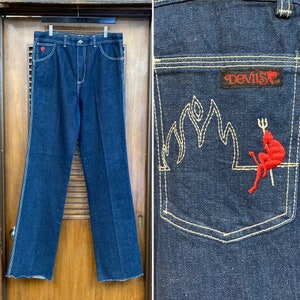 May include: A pair of dark blue denim jeans with a red heart on the left pocket and a red devil with a trident sitting on a flame embroidered on the back right pocket. The back pocket also has a red "Devil's" label.