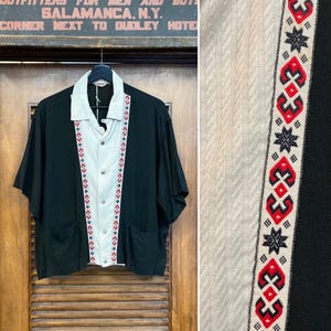 May include: Black short-sleeved shirt with a light gray center panel and a collar. The center panel has decorative red, white, and blue embroidered trim. The shirt has two front pockets and is hanging on a wooden door.