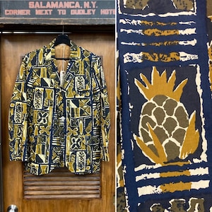 May include: A vintage blazer with a unique geometric pattern in shades of navy, olive green, and gold. The blazer is single-breasted with a two-button closure and a notched lapel. The fabric features a repeating design of abstract shapes and symbols. The blazer is hanging on a wooden door.