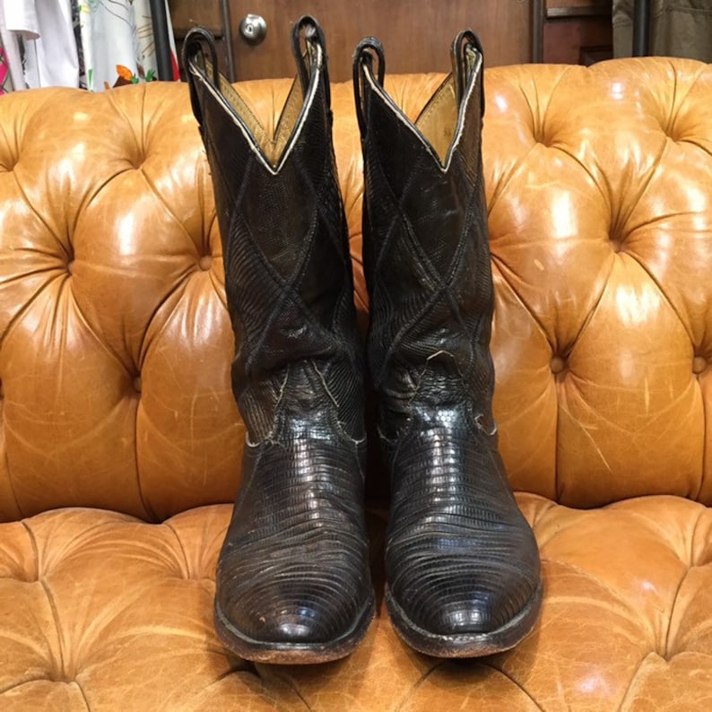 Pu&ograve; includere: Un paio di stivali da cowboy in pelle nera con punta a punta e un motivo testurizzato. Gli stivali sono su un divano in pelle marrone.