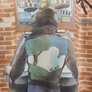 May include: A person wearing a brown leather jacket with a green, blue, and white design on the back. The jacket has the text "mik mik" written on the back. The person is standing in front of a brick wall and a vintage arcade game.
