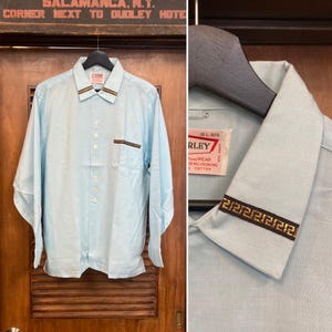 Puede incluir: Una camisa azul claro de manga larga con botones, bolsillo en el pecho y ribete decorativo en el cuello y el bolsillo. La camisa está colgada en una puerta de madera. La etiqueta dice "RLEY".