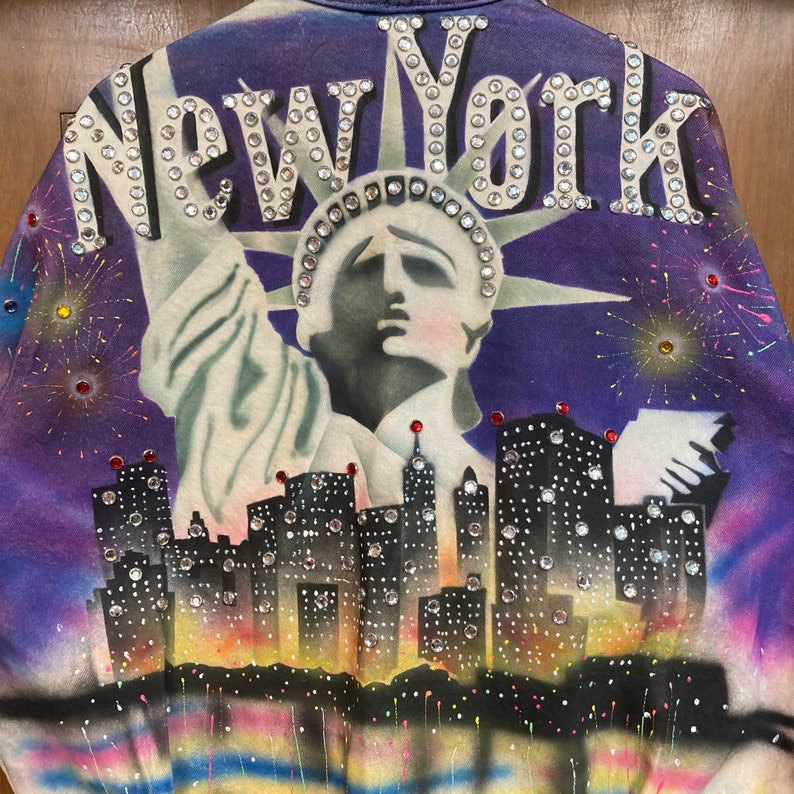 May include: A purple jacket with a rhinestone design of the Statue of Liberty and the words "New York". The background features a cityscape and fireworks.