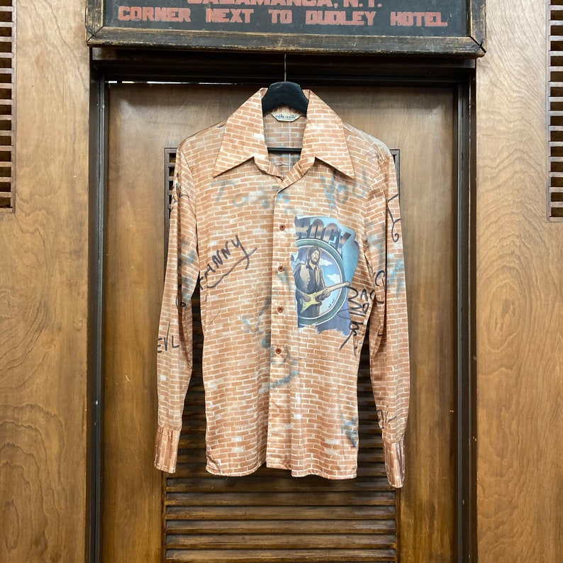 May include: A long-sleeved button-down shirt with a brick pattern and a graphic of a man playing a guitar. The shirt has the word "ZOOT" written on it.