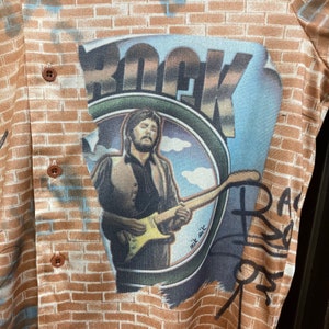 May include: A brown and tan patterned shirt with a graphic of a man playing a guitar in front of a brick wall. The graphic says "Rock" and "nik sik".