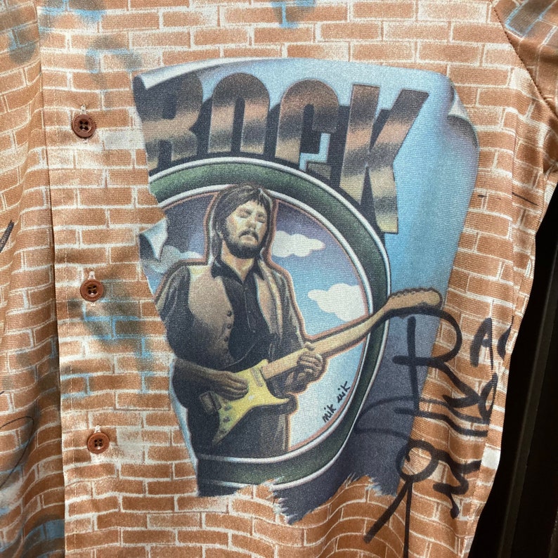 May include: A brown and tan patterned shirt with a graphic of a man playing a guitar in front of a brick wall. The graphic says "Rock" and "nik sik".