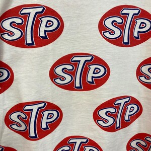 Peut inclure: Un t-shirt blanc avec un motif r&eacute;p&eacute;titif sur toute la surface du logo STP. Chaque logo se compose des lettres STP en blanc avec un contour bleu, centr&eacute;es dans un ovale rouge uni. Le tissu pr&eacute;sente une texture visible et l'impression est uniforme.