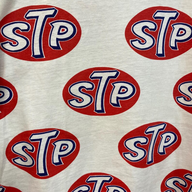 Peut inclure: Un t-shirt blanc avec un motif r&eacute;p&eacute;titif sur toute la surface du logo STP. Chaque logo se compose des lettres STP en blanc avec un contour bleu, centr&eacute;es dans un ovale rouge uni. Le tissu pr&eacute;sente une texture visible et l'impression est uniforme.