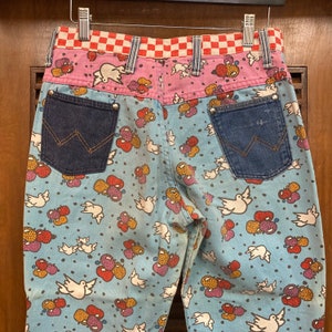 May include: Blue denim jeans with a white dove and colorful balloon pattern. The jeans have a pink and red checkered waistband and two blue denim pockets on the back.