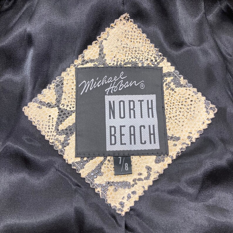 Puede incluir: Una etiqueta negra y beige con el texto "Michael Hoban" y "North Beach" en blanco. La etiqueta est&aacute; unida a una tela beige con un patr&oacute;n de piel de serpiente. La etiqueta tambi&eacute;n tiene el tama&ntilde;o "7/8" impreso en ella.