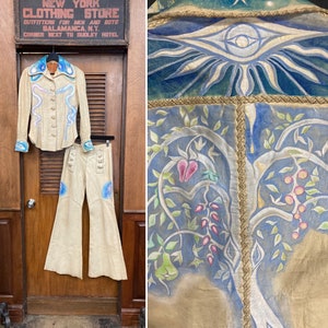 May include: A two-piece outfit made of light beige leather with a painted design. The top has a long sleeve, a collar, and a button-up front. The pants are flared and have a button-up front. The design on the outfit features a blue and white swirl pattern, a pink flower, and a green leaf.