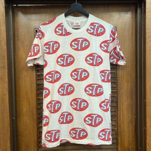 Peut inclure: Un t-shirt blanc &agrave; manches courtes avec un motif r&eacute;p&eacute;titif sur toute la surface compos&eacute; d'ovales rouges &agrave; contours bleus et du texte blanc STP &agrave; l'int&eacute;rieur. Le t-shirt a un col rond et est pr&eacute;sent&eacute; sur un cintre noir devant une porte en bois.