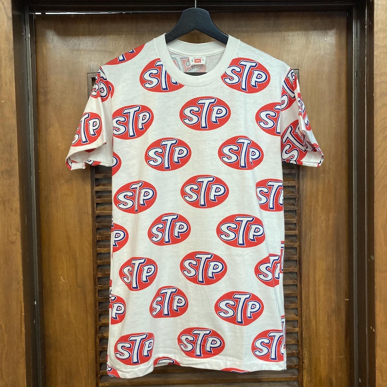 Peut inclure: Un t-shirt blanc &agrave; manches courtes avec un motif r&eacute;p&eacute;titif sur toute la surface compos&eacute; d'ovales rouges &agrave; contours bleus et du texte blanc STP &agrave; l'int&eacute;rieur. Le t-shirt a un col rond et est pr&eacute;sent&eacute; sur un cintre noir devant une porte en bois.