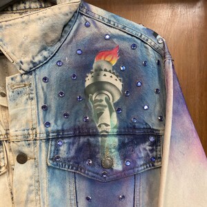 May include: A blue denim jacket with a tie-dye effect and a painted image of the Statue of Liberty on the left shoulder. The jacket is decorated with blue rhinestones.
