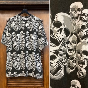 Puede incluir: Camiseta negra con un patr&oacute;n de calaveras blancas. Las calaveras est&aacute;n dispuestas en un patr&oacute;n aleatorio y son de diferentes tama&ntilde;os.