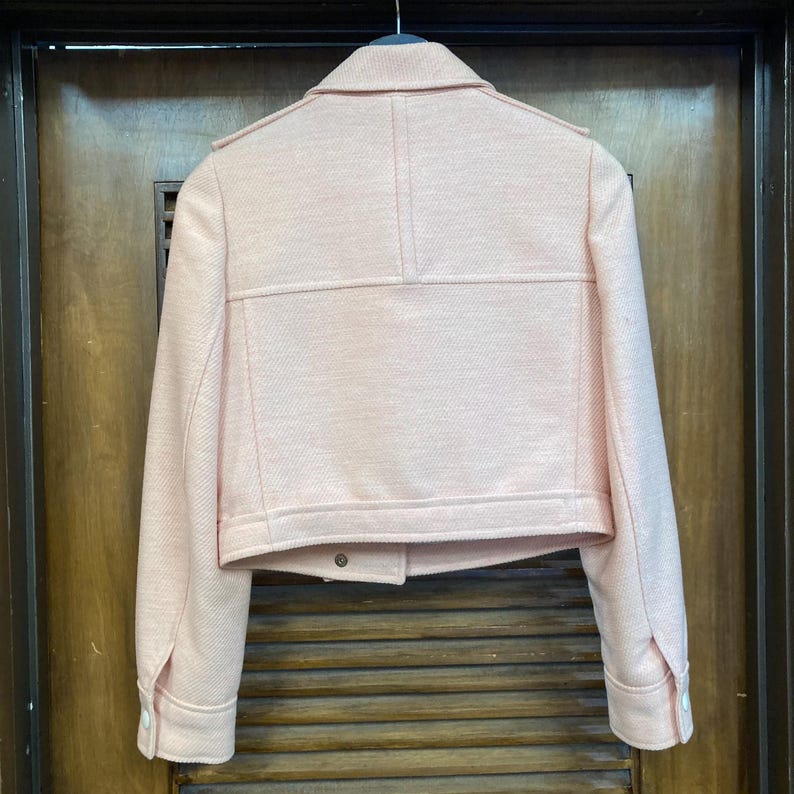 May include: A pink cropped jacket with a zipper closure and long sleeves. The jacket has a textured fabric and a slightly oversized fit.