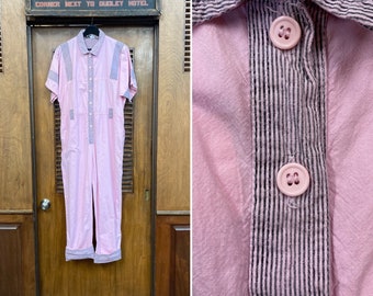 May include: A pink short-sleeved jumpsuit with a button-up front and black and white striped trim. The jumpsuit has a patch pocket on the left chest and two patch pockets on the front. The jumpsuit is hanging on a wooden door with a sign that reads "New York Clothing Store".