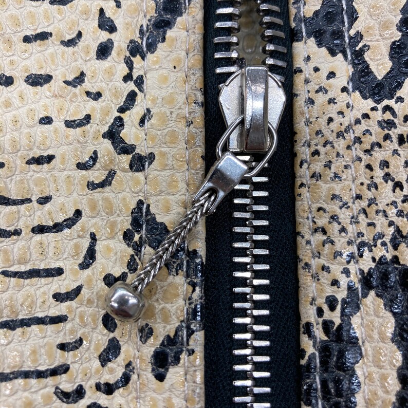 Puede incluir: Primer plano de una cremallera en una chaqueta de color beige y negro con estampado de piel de serpiente. La cremallera es plateada y tiene un tirador negro.