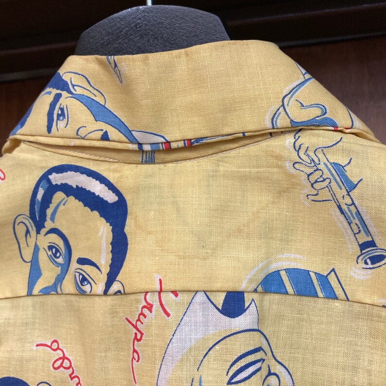 May include: A yellow button-down shirt with a colorful print of cartoon musicians. The shirt has a collar and a pocket on the left side. The text "Jungle" is visible on the shirt.