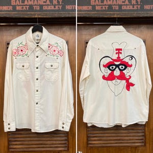 May include: Cream-colored button-down shirt with floral embroidery on the front and a graphic on the back. The front features two chest pockets and a row of buttons. The back has a red and black graphic design. The shirt is hanging on a wooden door.