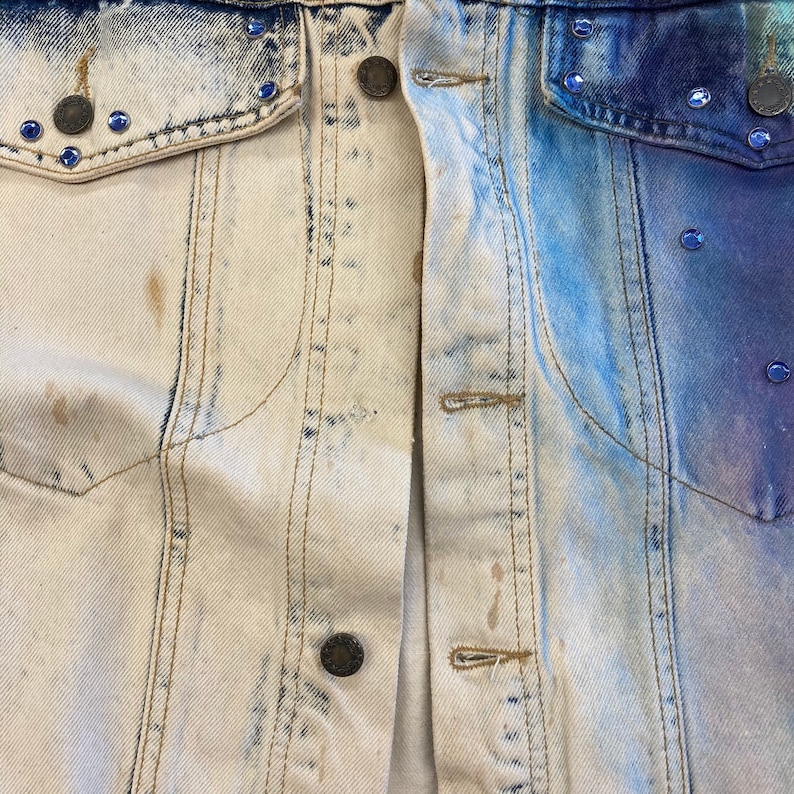 May include: A denim jacket with a light blue and white bleached design. The jacket has a button closure and two pockets on the front.