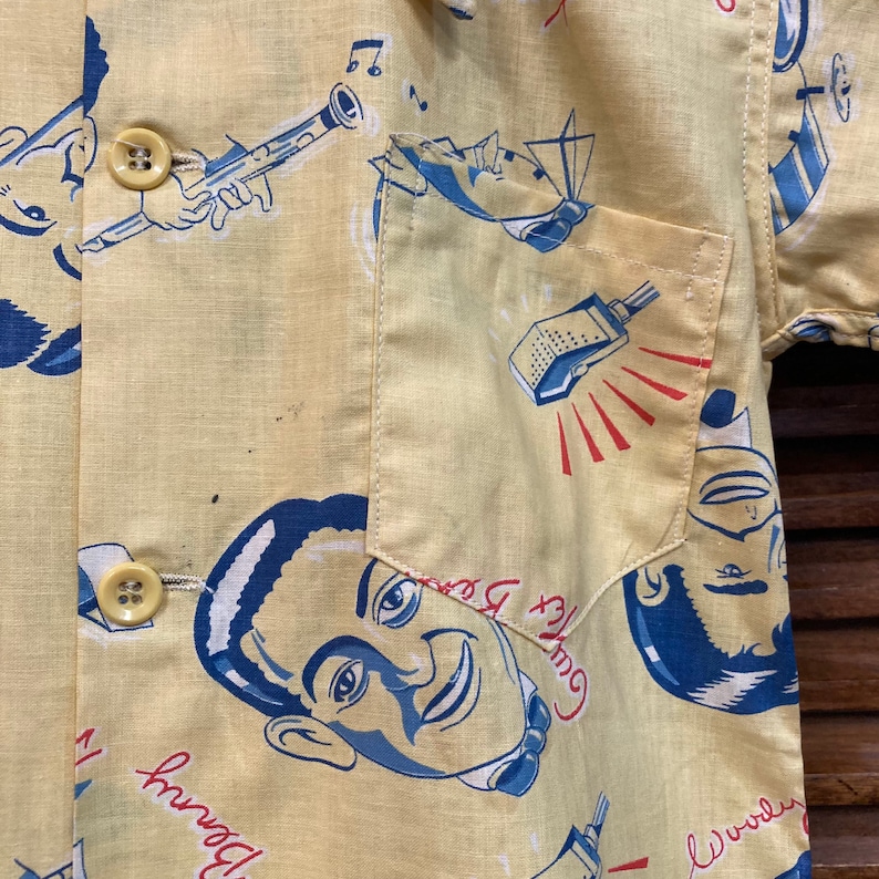 May include: A yellow short-sleeved shirt with a blue and red print of a man's face, a microphone, and musical notes. The shirt has a pocket on the left side and two buttons on the front.