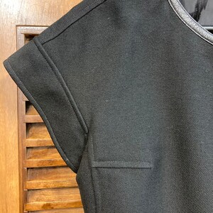 Puede incluir: Camiseta negra de manga corta con ribete de cuero negro alrededor del cuello. La camiseta tiene un tejido texturizado y un dise&ntilde;o sencillo.