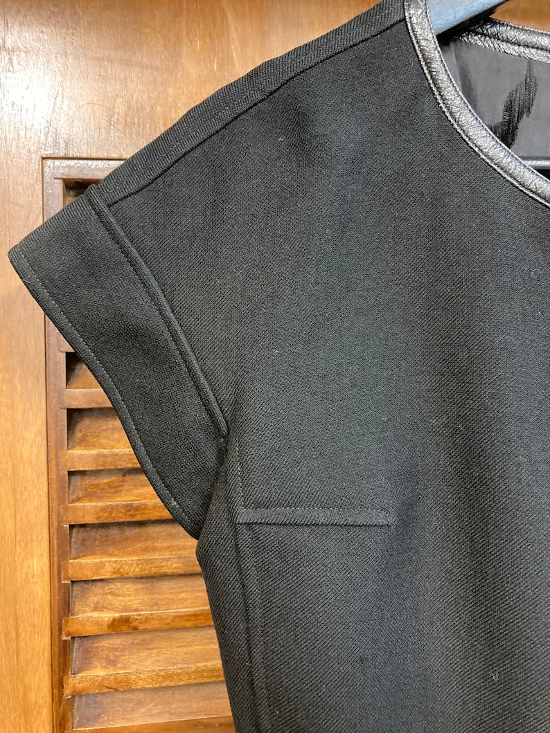 Puede incluir: Camiseta negra de manga corta con ribete de cuero negro alrededor del cuello. La camiseta tiene un tejido texturizado y un dise&ntilde;o sencillo.