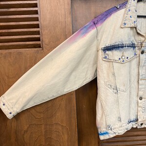 May include: A light blue denim jacket with a faded, tie-dye effect in shades of purple, pink, and blue. The jacket has a button closure and is decorated with rhinestones.