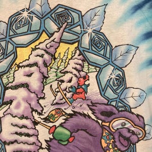 Puede incluir: Camiseta te&ntilde;ida con un dise&ntilde;o invernal. Un oso morado con gafas de esqu&iacute; desciende por una ladera nevada. El dise&ntilde;o incluye &aacute;rboles cubiertos de nieve, un esquiador y un borde floral decorativo en azul y blanco.