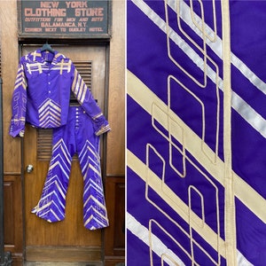 May include: A purple shirt and pants set with a geometric pattern in gold and white. The shirt has a button-down collar and long sleeves. The pants have a wide leg and a bell bottom style.
