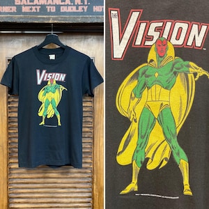 Könnte beinhalten: Schwarzes T-Shirt mit einem Vintage-Druck von The Vision aus Marvel Comics. The Vision ist in seinem klassischen grün-gelben Kostüm mit rotem Gesicht und einem gelben Diamanten auf der Stirn dargestellt. Der Text "The Vision" ist in Weiß über dem Druck gedruckt.