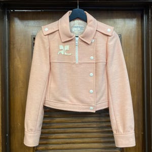 May include: A pink, cropped jacket with a zipper closure and white buttons. The jacket has a white logo on the front that says "Courreges Paris".