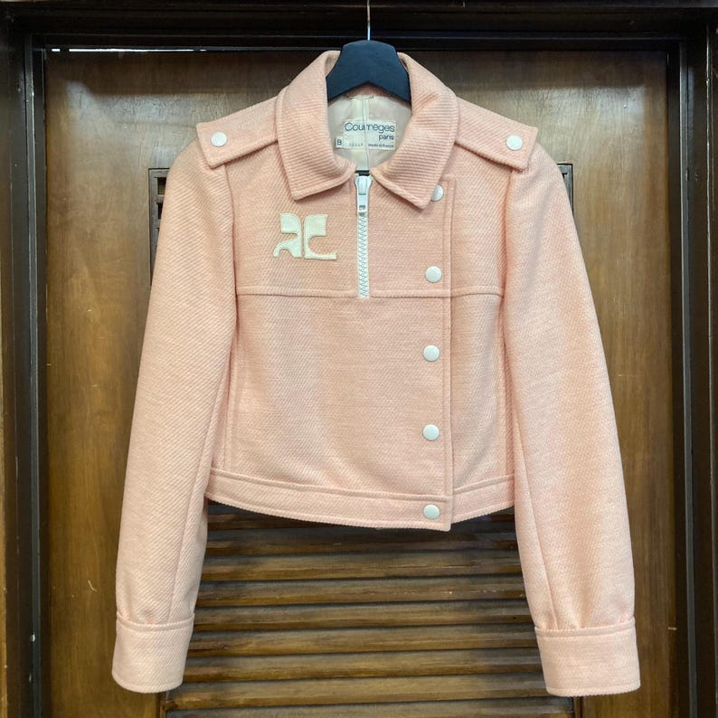 May include: A pink, cropped jacket with a zipper closure and white buttons. The jacket has a white logo on the front that says "Courreges Paris".