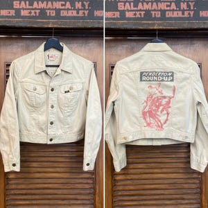 Puede incluir: Chaqueta vaquera beige con botones, bolsillos delanteros y cuello. La parte trasera de la chaqueta presenta un gráfico rojo y el texto "PENDLETON ROUND-UP". La chaqueta está colgada de una percha negra.