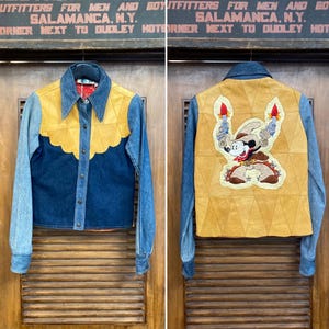 May include: A denim jacket with a yellow suede yoke and a navy blue front panel. The back features a tan suede panel with an embroidered cartoon character. The sleeves are a lighter blue denim. The jacket is button-down.