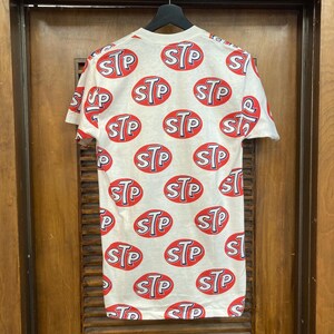Peut inclure: Un t-shirt blanc &agrave; manches courtes avec un motif r&eacute;p&eacute;titif sur toute la surface compos&eacute; de logos ovales rouges portant les lettres STP imprim&eacute;es en bleu et blanc. Le t-shirt est pr&eacute;sent&eacute; sur un cintre noir devant une porte en bois.