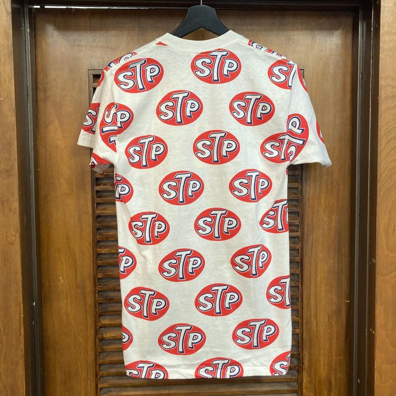Peut inclure: Un t-shirt blanc &agrave; manches courtes avec un motif r&eacute;p&eacute;titif sur toute la surface compos&eacute; de logos ovales rouges portant les lettres STP imprim&eacute;es en bleu et blanc. Le t-shirt est pr&eacute;sent&eacute; sur un cintre noir devant une porte en bois.